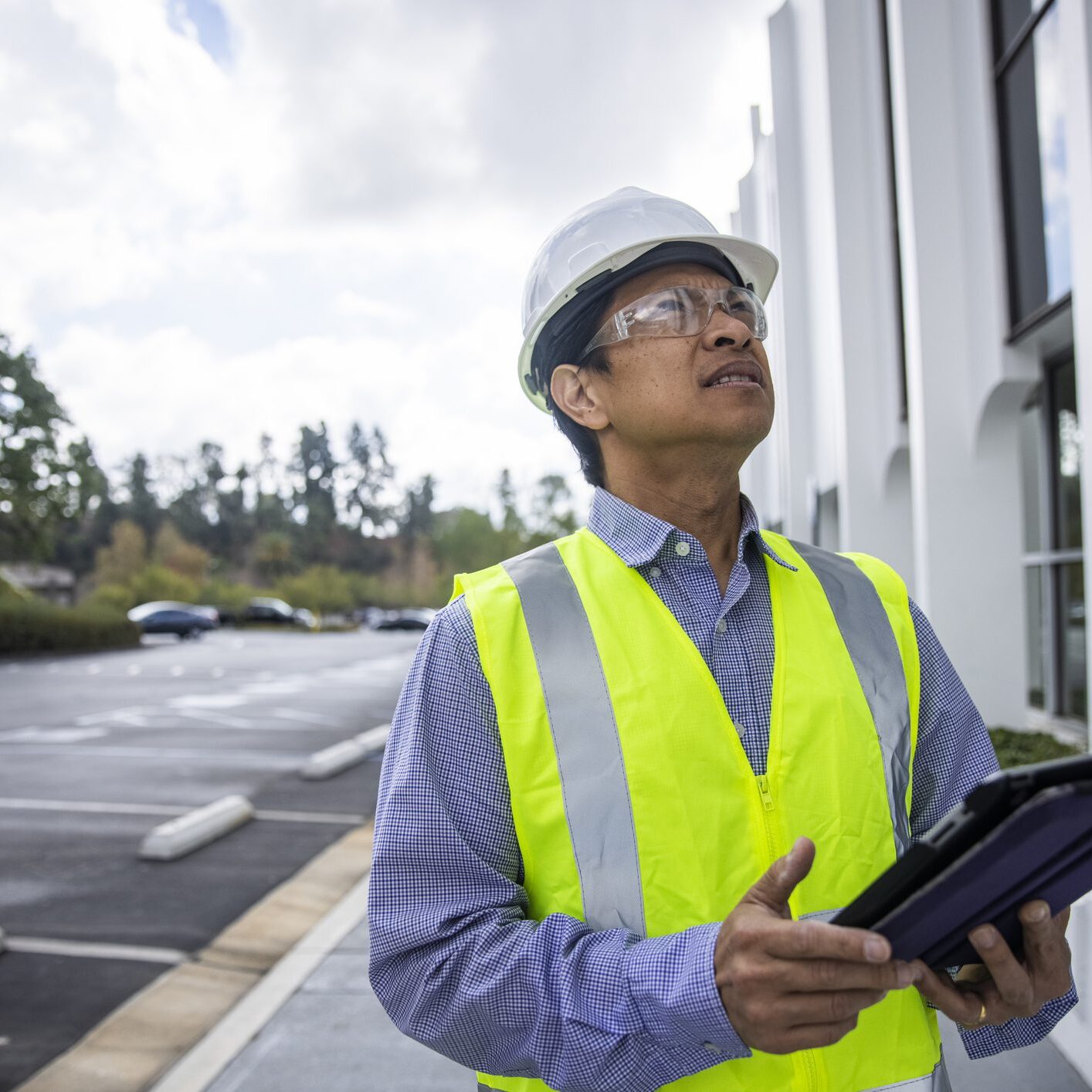 A mature Filipino man with a tablet and safety vest, helmet and glasses inspects a building and takes notes.